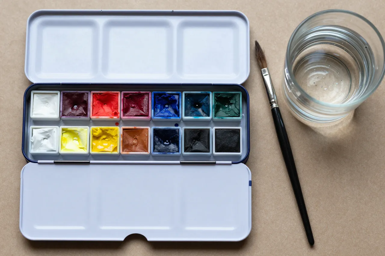 An open metal watercolor tin, one paintbrush, and a glass of water rest on a tan paper surface