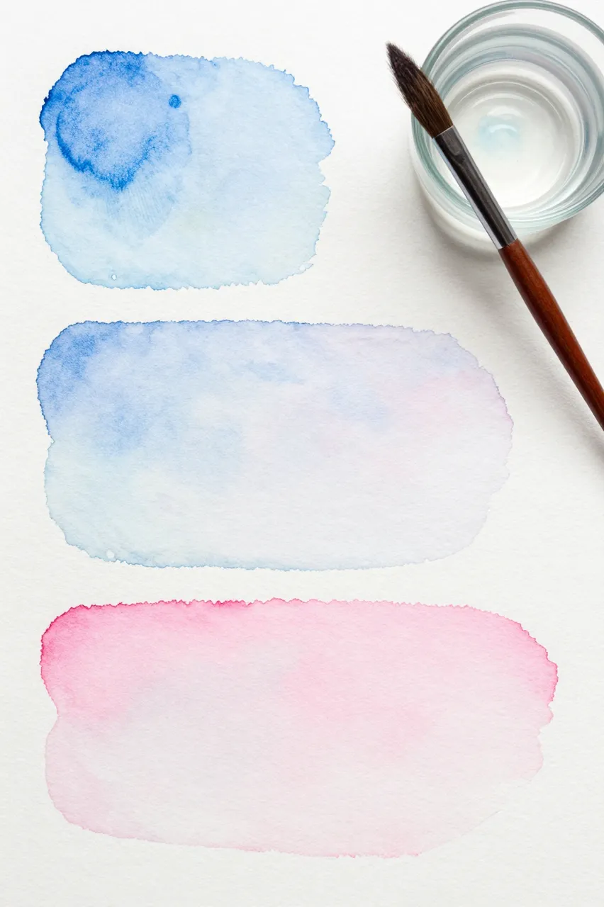 Flat lay of soft blue and pink watercolor washes, a round brush, and a glass water cup on textured paper
