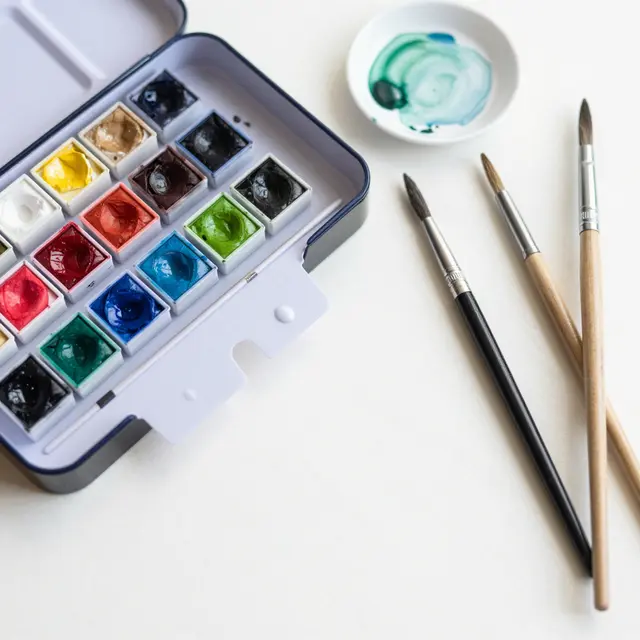 Top view of an open watercolor paint tin, mixing dish and paintbrushes arranged on a white surface