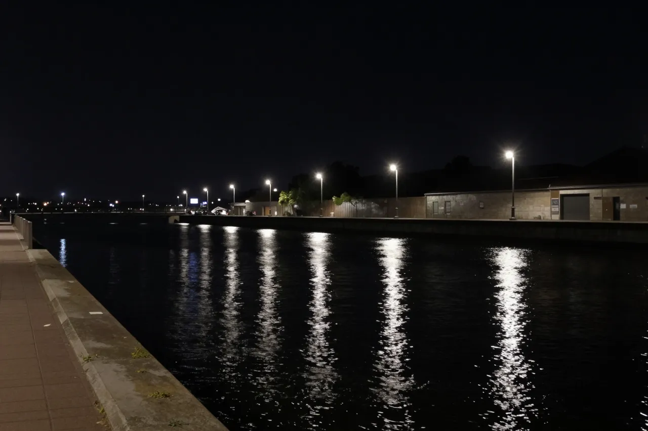 Lit street lamps cast vertical reflections on dark still water alongside an empty waterfront promenade at night