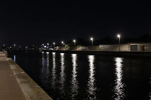 Lit street lamps cast vertical reflections on dark still water alongside an empty waterfront promenade at night