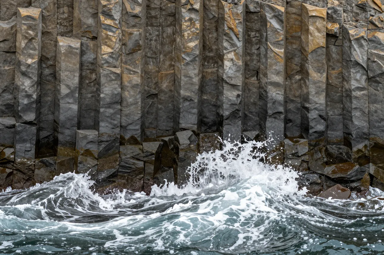 Turbulent ocean waves crash against tall vertical basalt columns along a rocky shoreline.