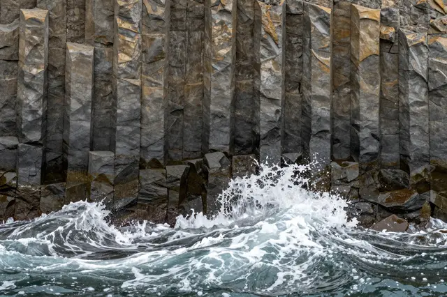 Turbulent ocean waves crash against tall vertical basalt columns along a rocky shoreline.