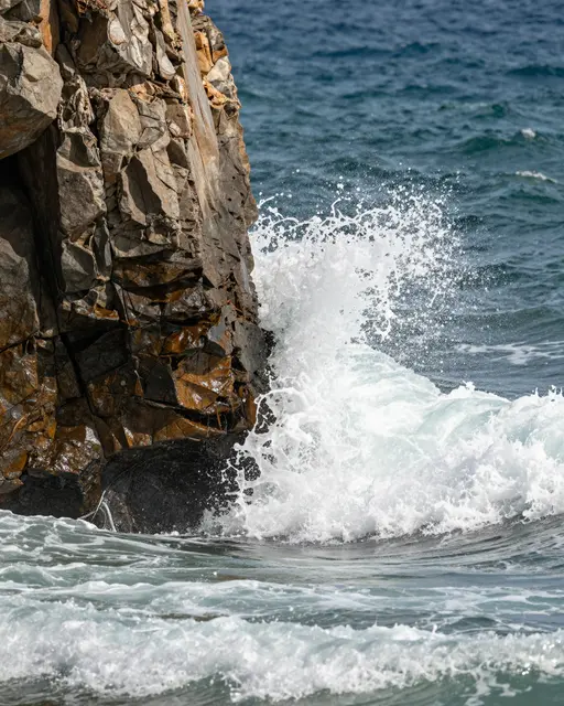 White surf crashes against a textured, weathered ocean rock pillar under deep blue sea water