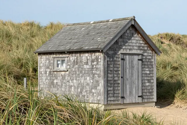 A weathered gray wood shingle beach hut sits on sandy ground surrounded by grassy coastal dunes under a pale blue sky.