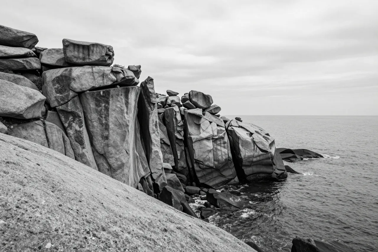 Black and white view of weathered granite cliffs dropping into calm sea under an overcast sky