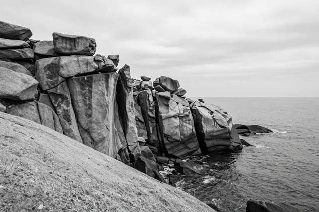 Black and white view of weathered granite cliffs dropping into calm sea under an overcast sky