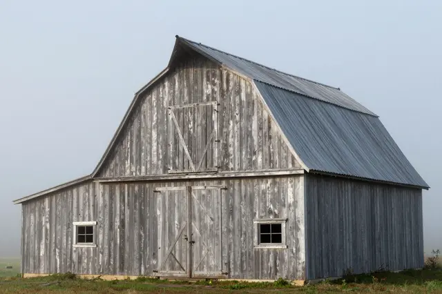 A weathered wooden barn stands in a rural field under soft, hazy morning fog.