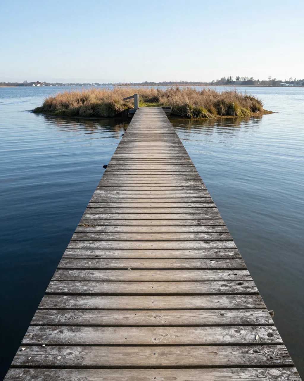 A weathered wooden jetty stretches across calm estuary water to a small grassy reed-covered islet under clear pale sky