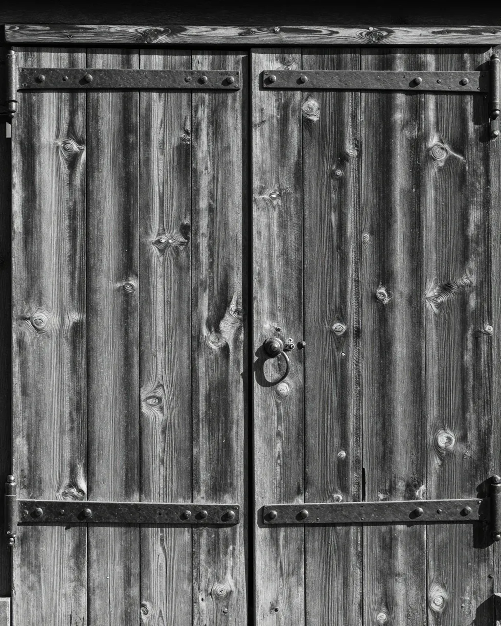 Black and white close-up of weathered wooden barn doors with iron strap hinges lit by side light