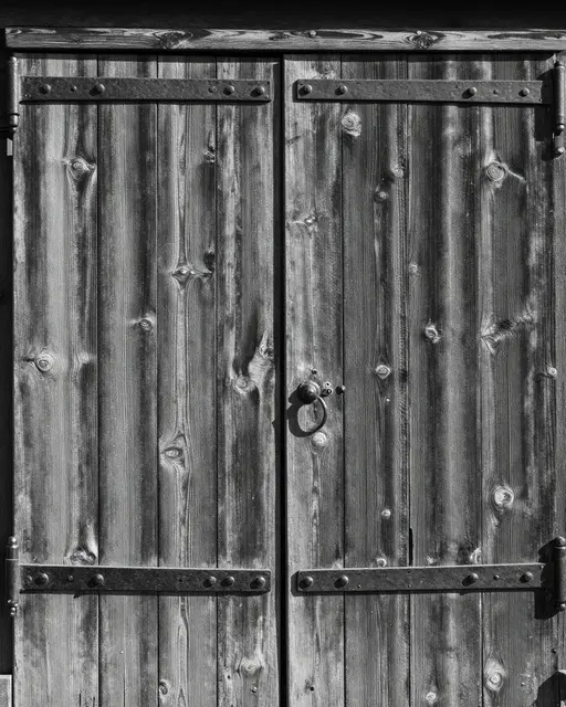 Black and white close-up of weathered wooden barn doors with iron strap hinges lit by side light