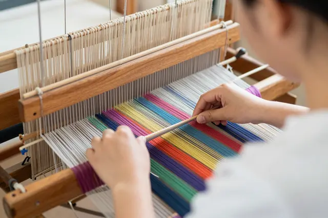 A weaver viewed from behind manipulates a wooden shuttle on a loom strung with colorful threads.