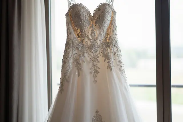 Beaded lace wedding dress hangs in soft natural light next to a sheer curtained window