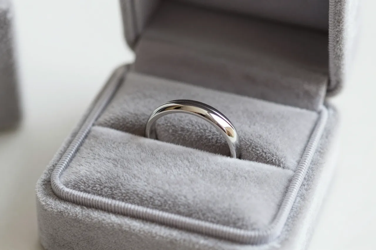 A polished silver wedding band rests inside an open gray velvet ring box on a light neutral surface.