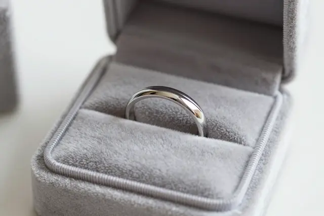 A polished silver wedding band rests inside an open gray velvet ring box on a light neutral surface.