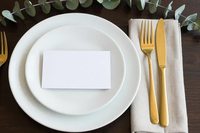 Flat lay of a wedding place setting with white stacked plates, gold cutlery, a blank card, linen napkin and eucalyptus atop