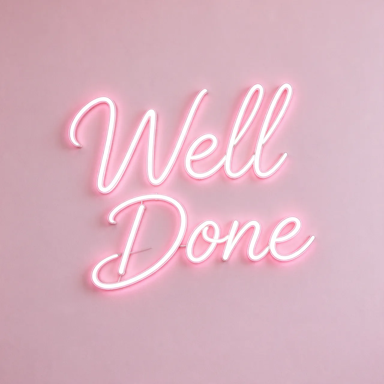 Glowing white neon script sign reading "Well Done" mounted on a solid pale pink wall