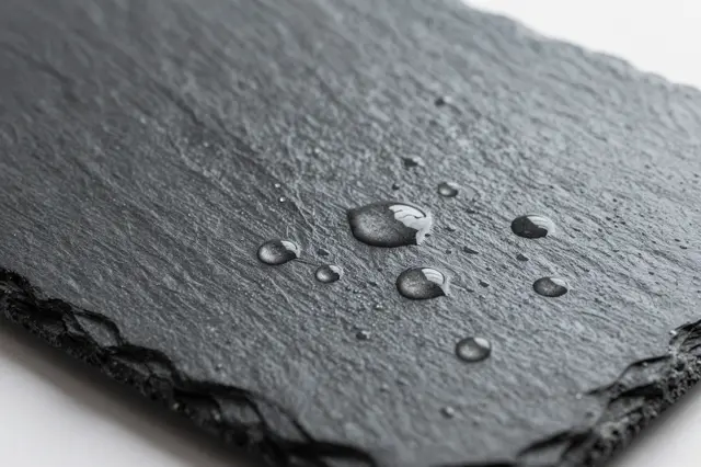 Close-up of a rough dark slate surface dotted with clear water droplets, set against a plain white background.