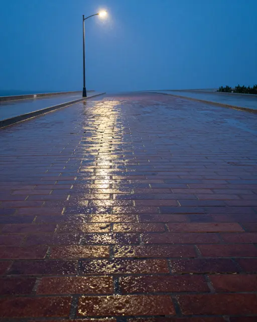 Lit streetlamp casts a golden reflection on rain-soaked brick pavement at blue hour