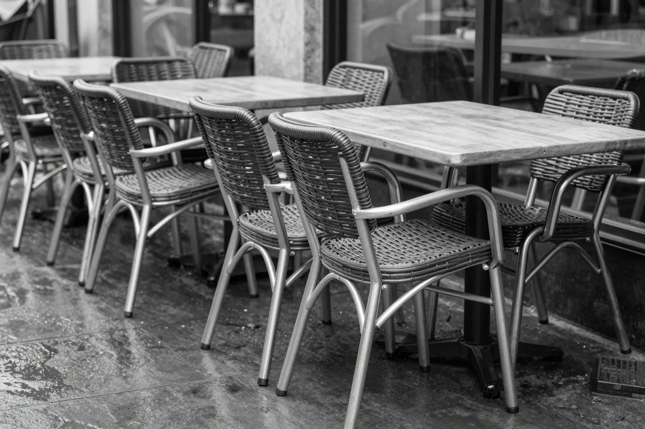 Stacked bistro chairs sit atop tables on a rain-dampened empty cafe terrace in black and white
