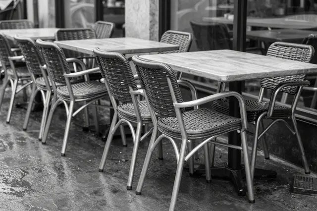 Stacked bistro chairs sit atop tables on a rain-dampened empty cafe terrace in black and white