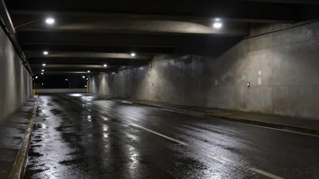 Dark empty concrete underpass with reflective wet asphalt pavement lit by overhead lights at night