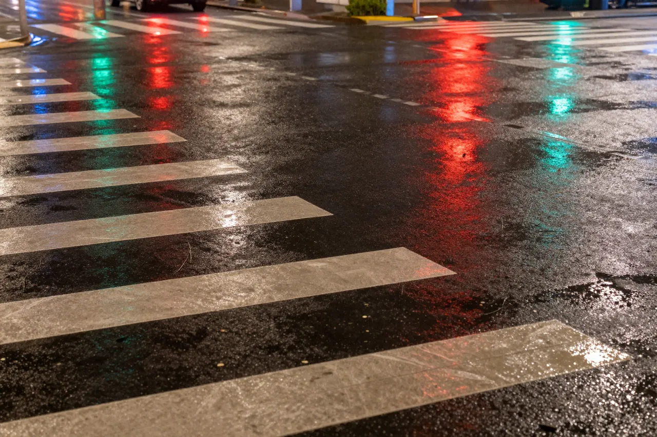 Wet asphalt crosswalk at night shows glowing red and green traffic light reflections from rain.