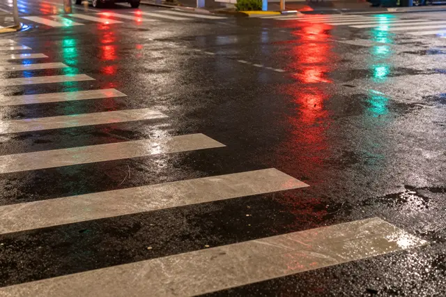 Wet asphalt crosswalk at night shows glowing red and green traffic light reflections from rain.