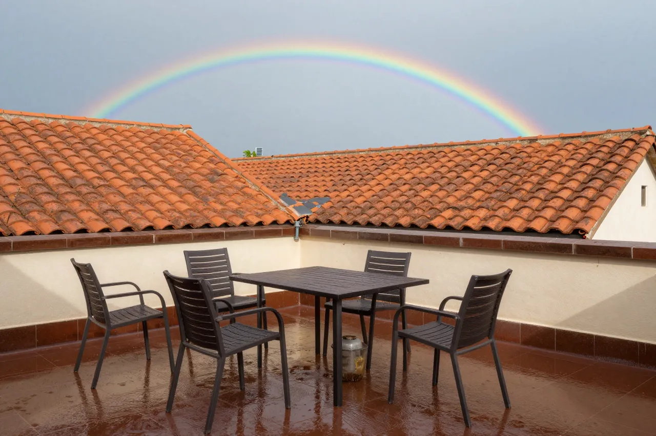 Wet tiled rooftop terrace with patio furniture, terracotta roofs and a rainbow after summer rain