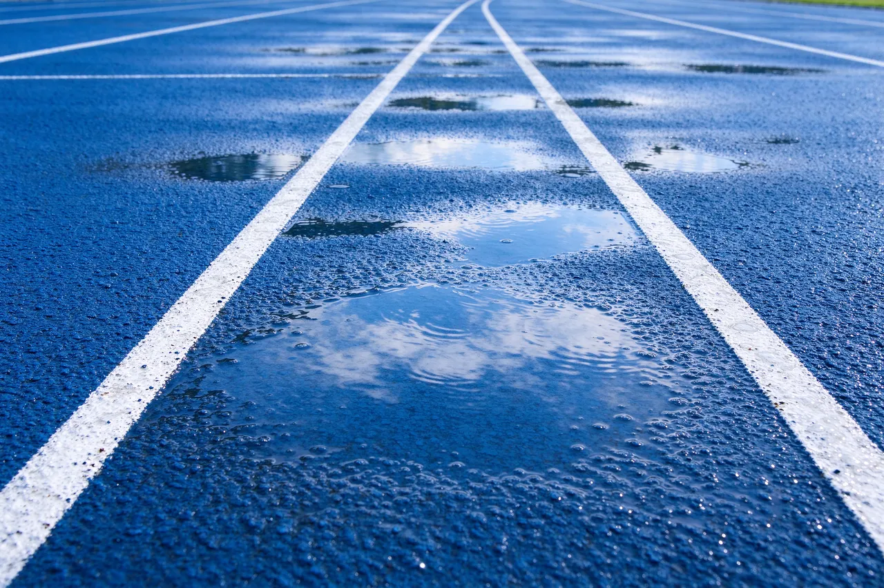 Close-up of a blue running track with white lane lines and rain puddles reflecting the sky