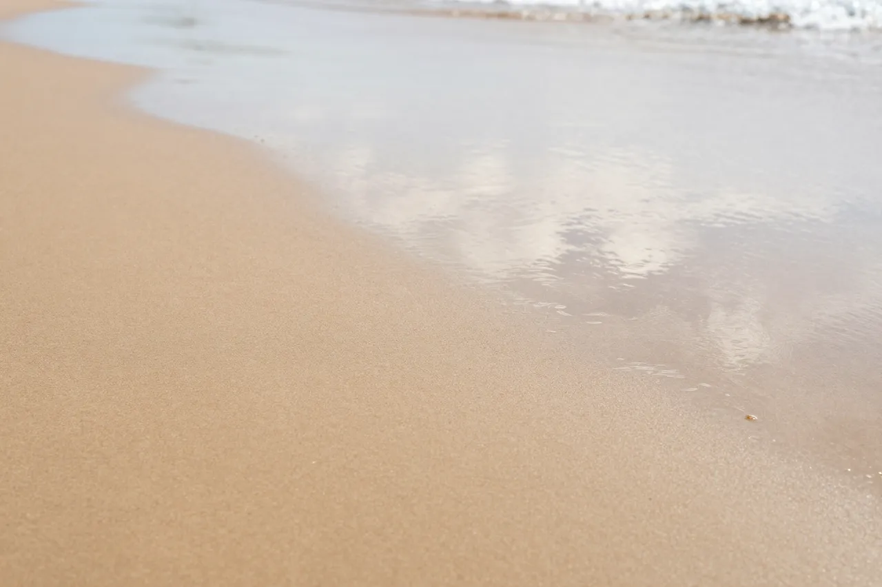 Gentle shallow ocean water laps onto smooth wet beach sand with soft sky reflections.