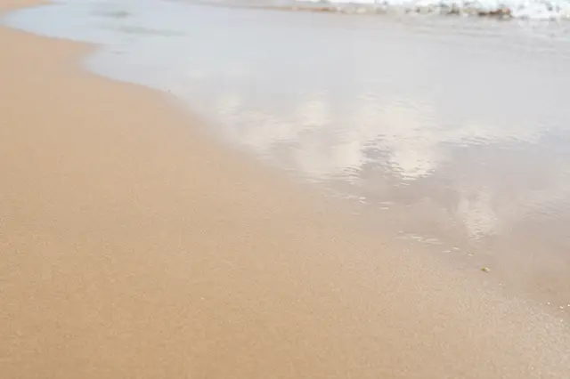 Gentle shallow ocean water laps onto smooth wet beach sand with soft sky reflections.