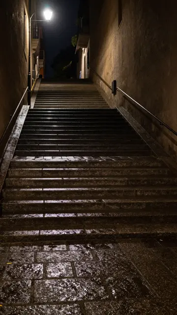 Illuminated steep wet stone staircase between building walls under a glowing street lamp after dark