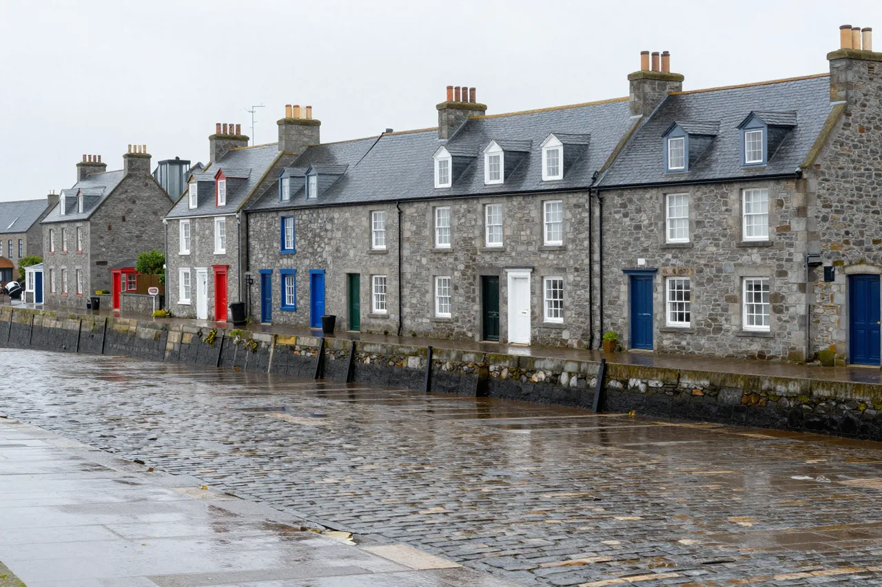 Rainy overcast harbor front with a row of gray stone houses lining a wet cobbled quay