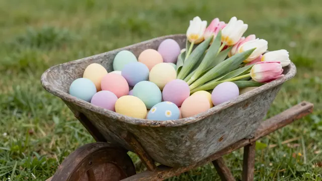 A rustic vintage wheelbarrow holds pastel Easter eggs and fresh tulips on a grass lawn.