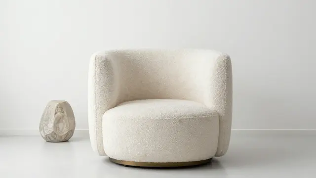 A rounded cream boucle armchair sits beside a small stone sculpture in an all-white minimalist room