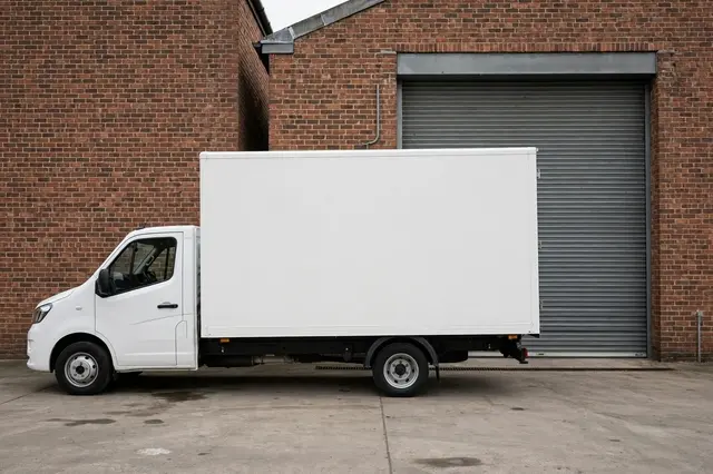 Side view of a plain unmarked white box truck parked outside a brick warehouse with a closed roller door