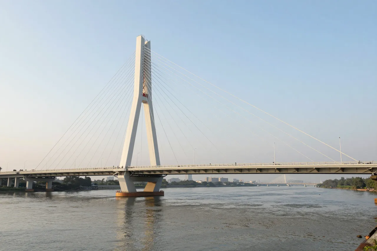 A tall white cable-stayed bridge spans a wide calm river under a clear pale morning sky