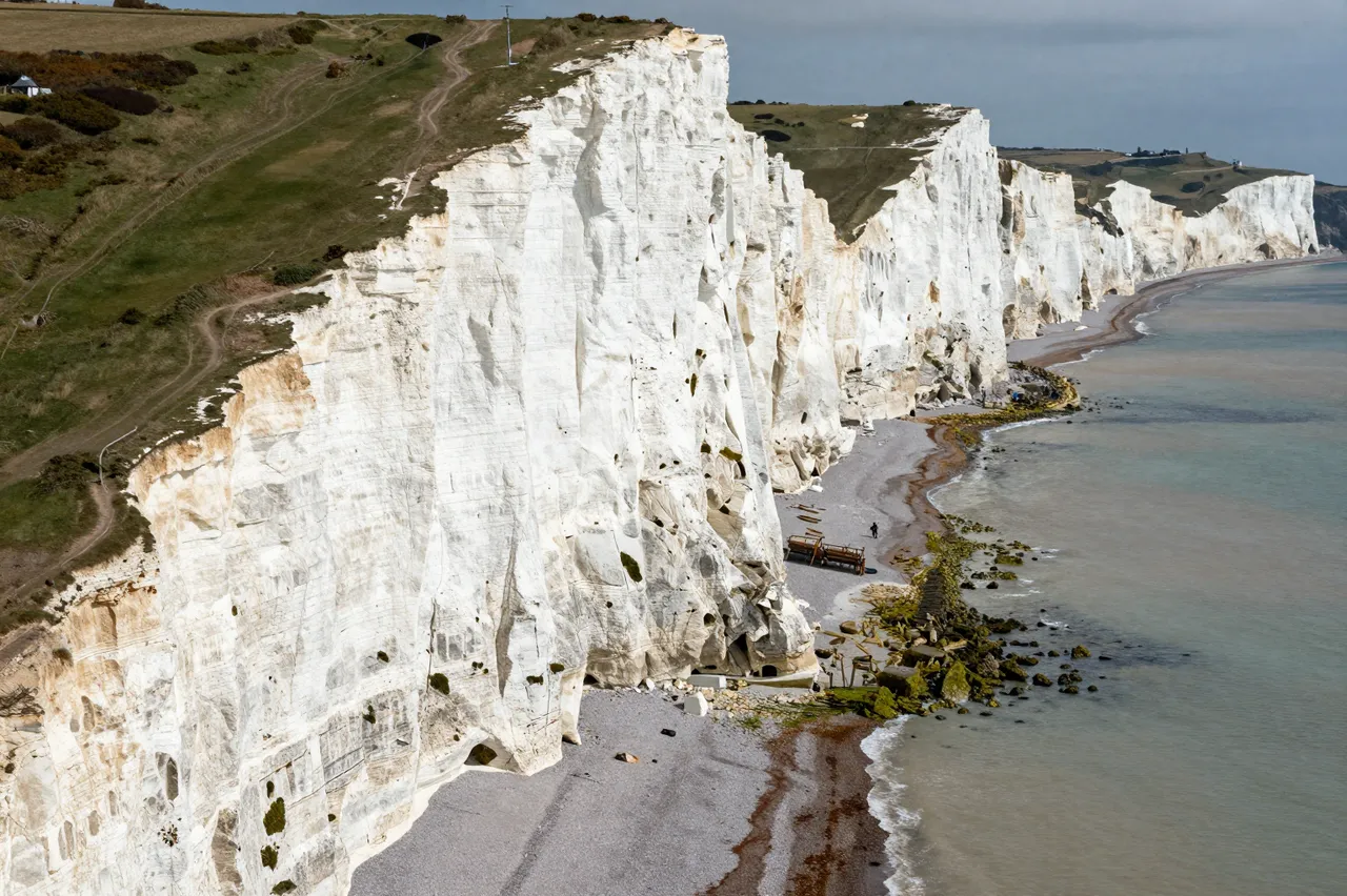 Aerial view of tall white chalk sea cliffs above a narrow pebbly beach along a calm coastal shoreline