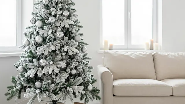 Flocked Christmas tree with silver baubles stands next to a cream sofa and candles on windowsills in a bright neutral living