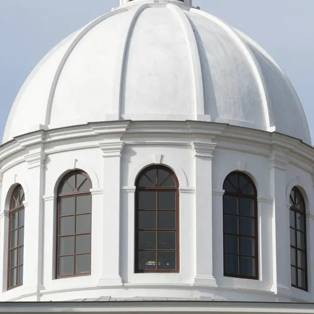 Exterior upward view of a smooth white church dome with arched brown-framed windows at its base against pale sky