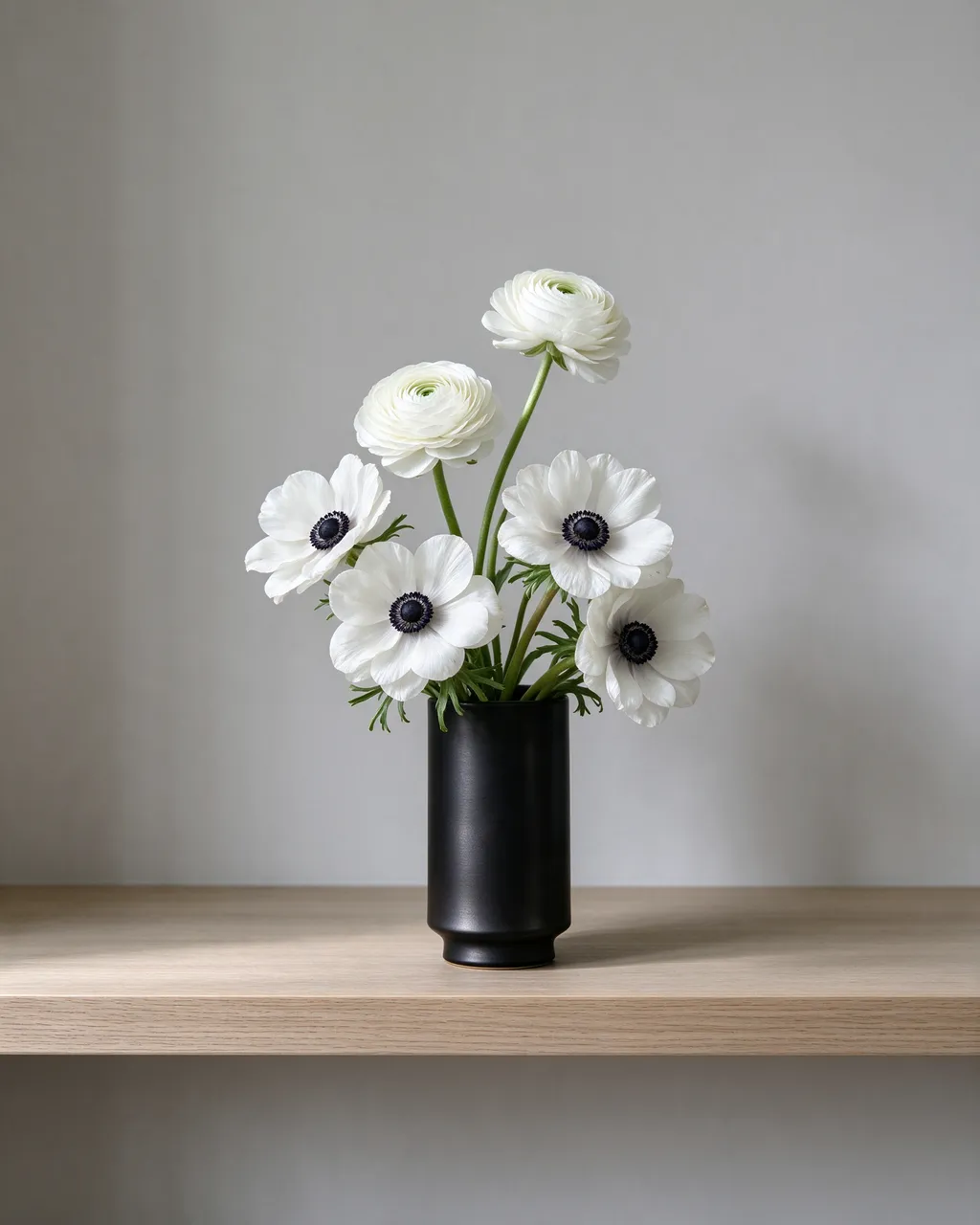 White ranunculus and anemone flowers sit in a matte black vase on a pale oak shelf against a light gray wall