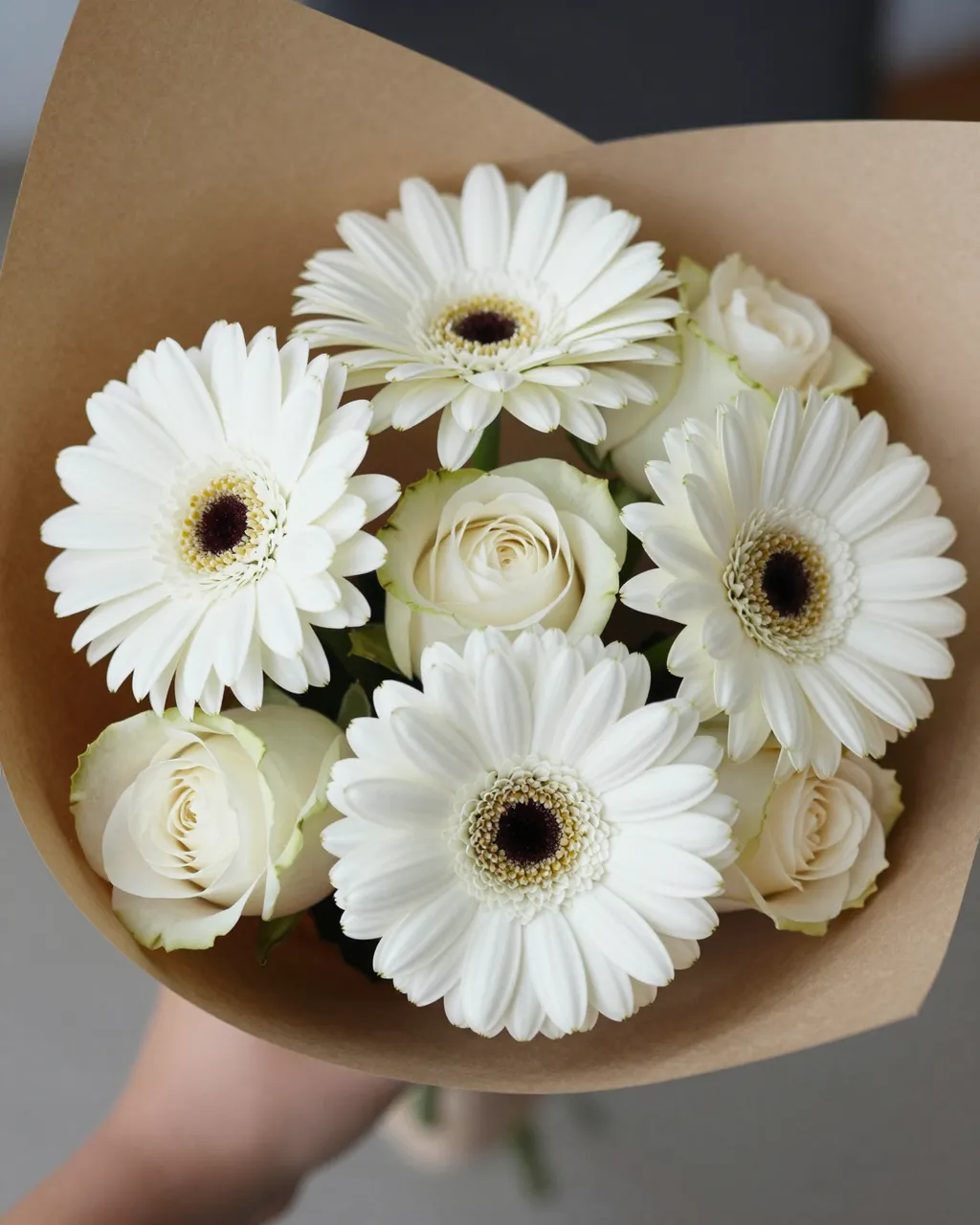 A close-up of a bouquet of white gerberas and roses wrapped in brown kraft paper held by a partially visible arm