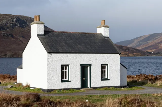 A whitewashed stone cottage sits on the shore of a Scottish highland loch beneath rolling moorland hills.