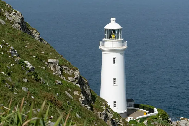 A tall white lighthouse stands on a rocky green grassy cliff overlooking a calm deep blue sea.