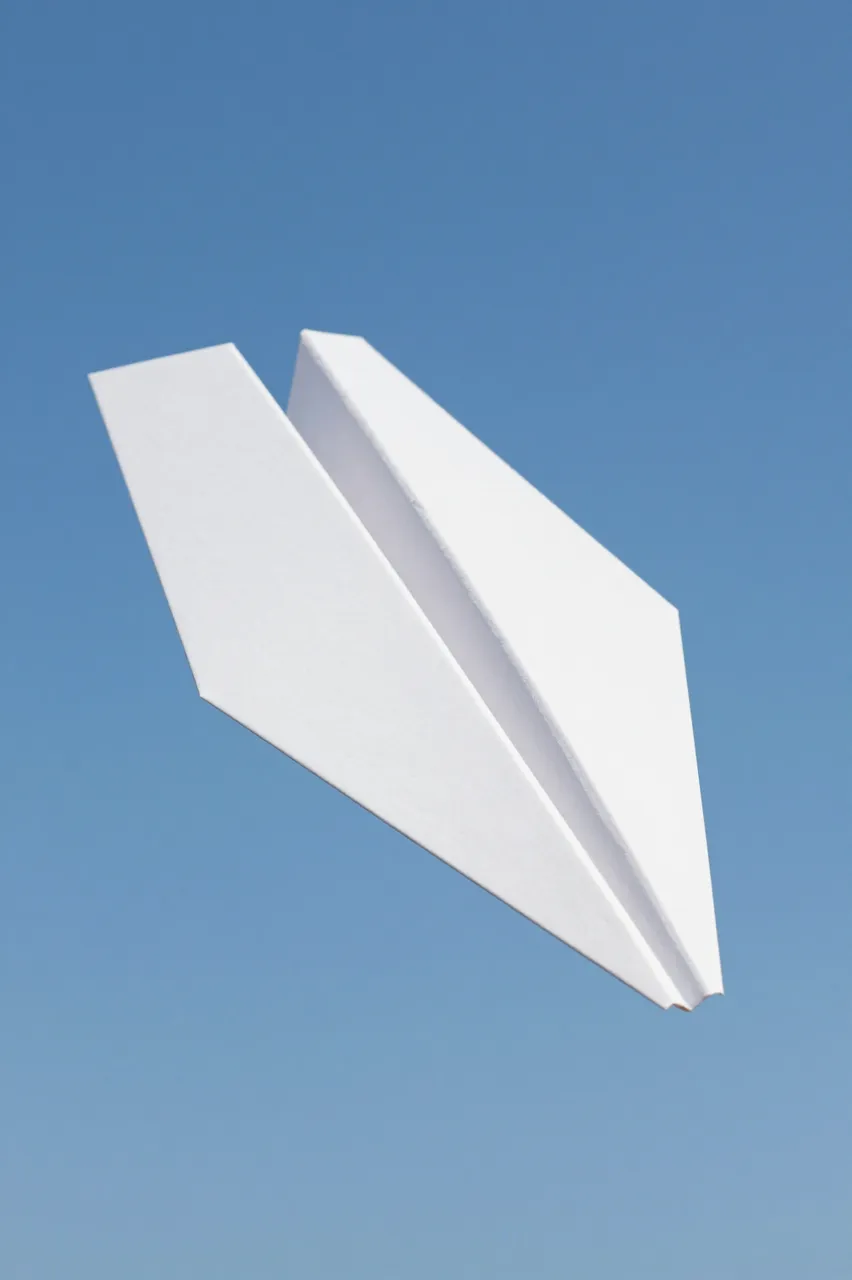 A white folded paper airplane glides against a smooth clear blue sky from a low angle