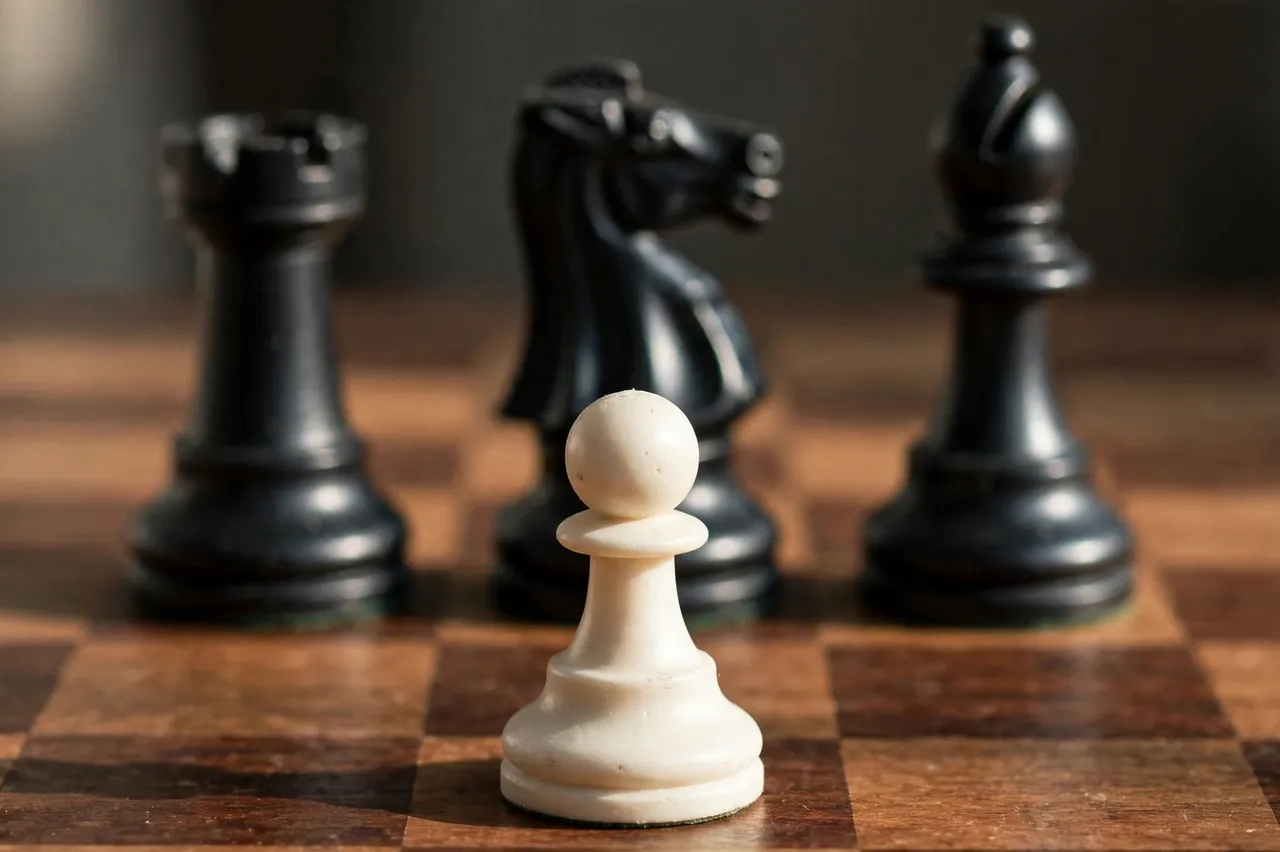 A close-up of a white chess pawn positioned in front of three out-of-focus black pieces on a wooden chessboard.