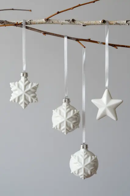 Glossy white porcelain snowflake and star ornaments hang at different heights from a bare birch branch against a pale gray