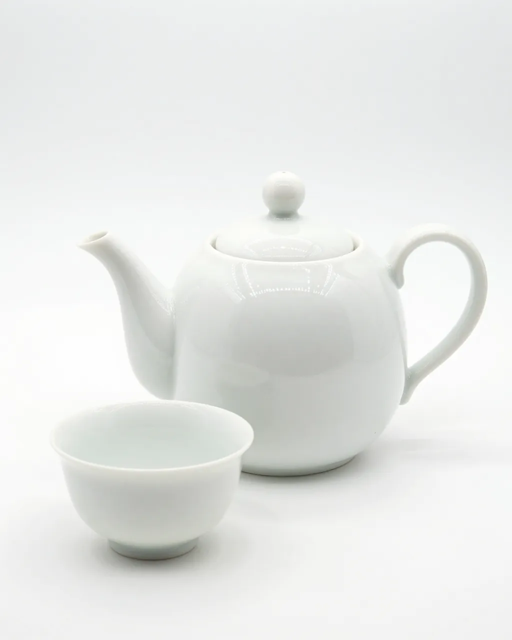 A glossy round white porcelain teapot sits next to a matching small teacup on a plain white background.