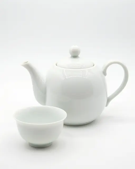 A glossy round white porcelain teapot sits next to a matching small teacup on a plain white background.